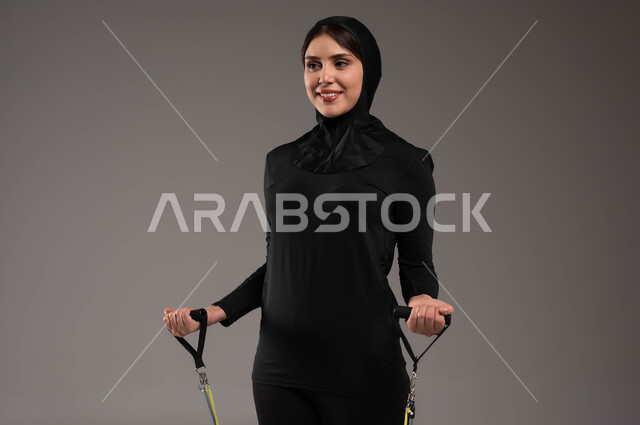 التمرن مع قبضة اليد المطاطية و المحافظةعلى أسلوب حياة صحي، تقوية عضلات الجسم والحفاظ على اللياقة، تمارين اليدين والصدرالمتوسع وحبل المقاومة، نشاط بدني للعناية بالصحة الجسدية، محاولة انقاص الوزن، بورتريه مقرب لشابة عربية خليجية سعودية محجبة، خلفية رمادية