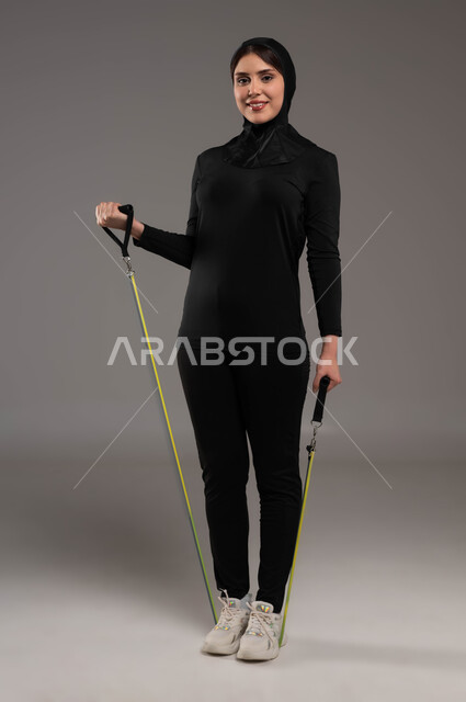 النظر إلى الكاميرا بإيماءات السعادة والنشاط، تمارين اليدين والصدر المتوسع وحبل المقاومة، محاولة انقاص الوزن، بورتريه لشابة عربية خليجية سعودية محجبة تقوم بالتمرين مع قبضة اليد المطاطية، أدوات ومعدات رياضية، أسلوب حياة صحي، صورة لكامل الجسم، خلفية رمادية