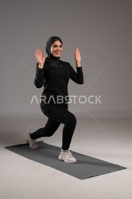إيماءات السعادة والنشاط، انشطة ورياضات لتقوية العضلات ونحت الجسم، أسلوب حياة صحي، بورتريه لشابة عربية خليجية سعودية  محجبة ترتدي زي رياضي تقوم بممارسة تمارين رياضية سكوات، نشاط بدني للمحافظة على صحة و لياقة الجسم، صورة لكامل الجسم، خلفية رمادية