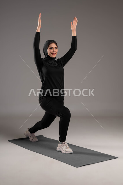 ممارسة تمارين رياضية سكوات، انشطة ورياضات لتقوية العضلات ونحت الجسم، إيماءات السعادة والنشاط، أسلوب حياة صحي، بورتريه لشابة عربية خليجية سعودية  محجبة ترتدي زي رياضي، نشاط بدني للمحافظة على صحة و لياقة الجسم، صورة لكامل الجسم، خلفية رمادية