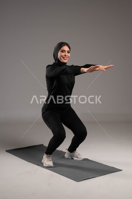 النظر إلى الكاميرا بإيماءات السعادة، انشطة ورياضات لتقوية العضلات ونحت الجسم، أسلوب حياة صحي، بورتريه لشابة عربية خليجية سعودية  محجبة ترتدي زي رياضي تقوم بممارسة تمارين رياضية سكوات، نشاط بدني للمحافظة على صحة و لياقة الجسم، صورة لكامل الجسم، خلفية رمادية