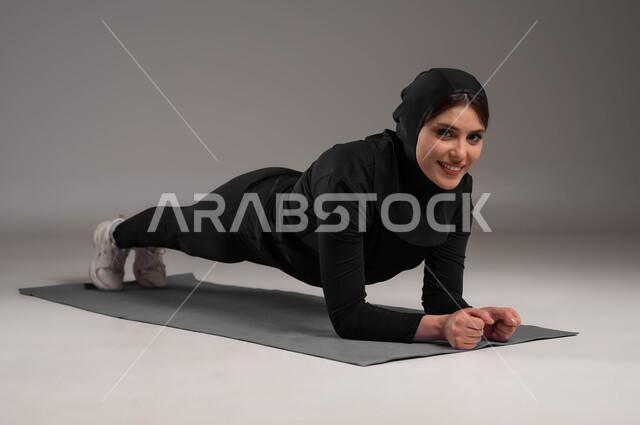 ممارسة تمارين رياضية للمحافظة على التوازن الجسدي، أسلوب حياة صحي، انشطة ورياضات لتقوية العضلات ونحت الجسم، بورتريه لشابة عربية خليجية سعودية  محجبة ترتدي زي رياضي، نشاط بدني للمحافظة على صحة و لياقة الجسم، صورة لكامل الجسم، خلفية رمادية