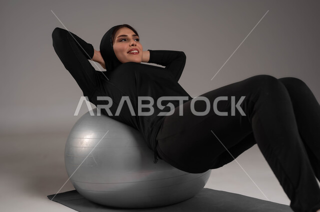 توفير أجهزة و معدات رياضية، ايماءات السعادة والنشاط، مفهوم الرشاقة واللياقة، انشطة رياضية لتقوية العضلات ونحت الجسم، أسلوب حياة صحي، بورتريه لشابة عربية خليجية سعودية ترتدي زي رياضي تقوم بممارسة تمارين باستخدام كرة البيلاتيس، صورة لكامل الجسم، خلفية رمادية