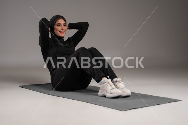 ممارسة تمرينات المقاومة لشد المعدة، سلوب حياة صحي، مفهوم الرشاقة واللياقة، انشطة رياضية لتقوية العضلات ونحت الجسم، المحافظة على التوازن الجسدي، ايماءات السعادة والنشاط، بورتريه لشابة عربية خليجية سعودية ترتدي زي رياضي أسود، صورة لكامل الجسم، خلفية رمادية