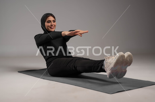 ممارسة التمرينات الأرضية لشد الجسد، اسلوب حياة صحي، مفهوم الرشاقة واللياقة، انشطة رياضية لتقوية العضلات ونحت الجسم، المحافظة على التوازن الجسدي، بورتريه لشابة عربية خليجية سعودية محجبة ترتدي زي رياضي أسود، خلفية رمادية