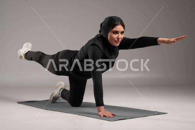 ممارسة التمرينات الأرضية لشد الجسد، اسلوب حياة صحي، مفهوم الرشاقة واللياقة، انشطة رياضية لتقوية العضلات ونحت الجسم، المحافظة على التوازن الجسدي، بورتريه لشابة عربية خليجية سعودية محجبة ترتدي زي رياضي أسود، خلفية رمادية
