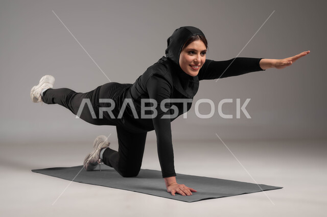 A healthy and beneficial lifestyle, practicing floor exercises to tighten the body, the concept of agility and fitness, sports activities to strengthen the muscles and sculpt the body, maintaining physical balance, a portrait of a veiled Saudi Gulf Arab woman wearing a black sports uniform, gray background