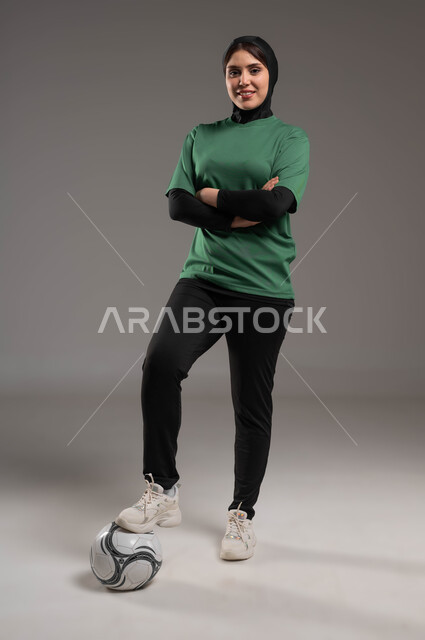 الاستعداد لبدء المباراة، المحافظة على أسلوب حياة صحي، ممارسة هواية اللعب بكرة القدم، بورتريه للاعبة عربية سعودية خليجية محجبة مبتسمة ترتدي الملابس الرياضية تقف بإيدي متقاطعة وتنظر إلى الكاميرا بتعابير السرور، صورة الجسم بالكامل، خلفية رمادية.