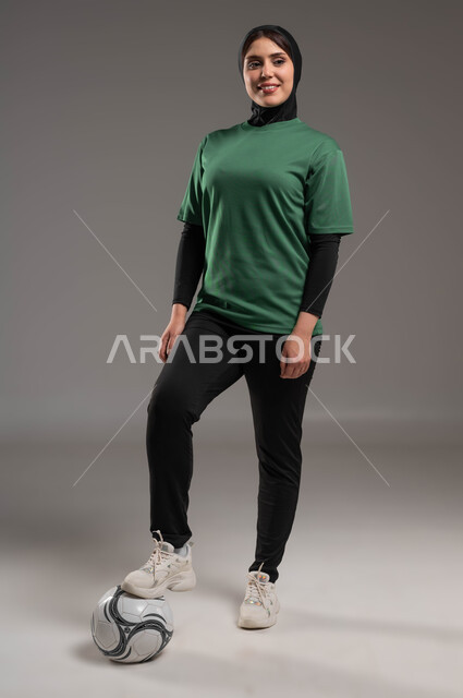 المحافظة على أسلوب حياة صحي، الاستعداد لبدء المباراة، ممارسة هواية اللعب بكرة القدم، بورتريه للاعبة عربية سعودية خليجية محجبة مبتسمة ترتدي الملابس الرياضية تنظر إلى الكاميرا بتعابير السرور، احتراف الرياضات الجماعية، صورة الجسم بالكامل، خلفية رمادية.