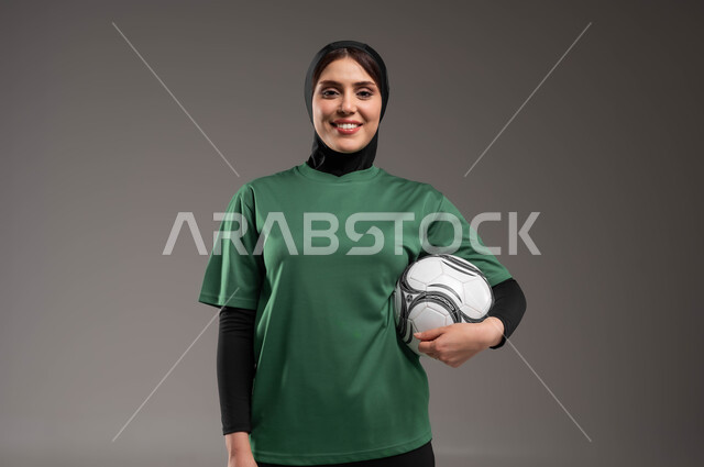 الوقوف باستقامة والنظر إلى الكاميرا، المحافظة على أسلوب حياة صحي، الاستعداد لبدء المباراة، ممارسة هواية اللعب بكرة القدم، بورتريه للاعبة عربية سعودية خليجية ترتدي الملابس الرياضية تحمل بيدها الكرة بإيماءات السرور، احتراف الرياضات الجماعية، خلفية رمادية.