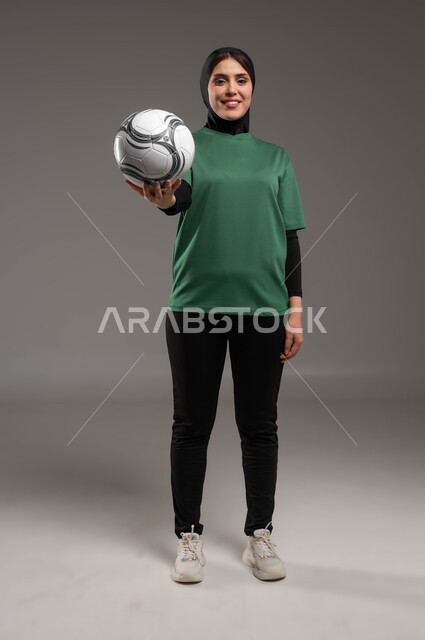 الوقوف باستقامة والنظر إلى الكاميرا، المحافظة على أسلوب حياة صحي، الاستعداد لبدء المباراة، ممارسة هواية اللعب بكرة القدم، بورتريه للاعبة عربية سعودية خليجية ترتدي الملابس الرياضية تحمل بيدها الكرة، احتراف الرياضات الجماعية، صورة الجسم بالكامل، خلفية رمادية.