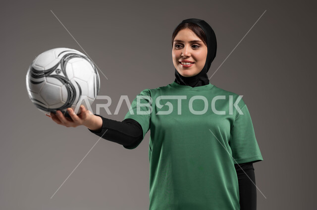 الاستمتاع بممارسة هواية اللعب بكرة القدم، احتراف الرياضات الجماعية، المحافظة على أسلوب حياة صحي، الاستعداد لبدء المباراة، بورتريه مقرب للاعبة عربية خليجية سعودية محجبة مبتسمة ترتدي الملابس الرياضية تحمل بيدها الكرة بإيماءات السرور، خلفية رمادية.