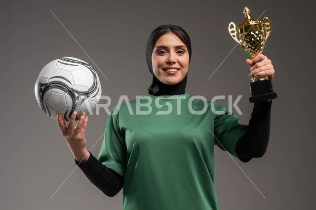 النظر إلى الكاميرا بإيماءات السعادة بالفوز والنصر، احتراف الرياضات الجماعية، المحافظة على نمط حياة صحي، بورتريه مقرب للاعبة عربية خليجية سعودية محجبة مبتسمة ترتدي الملابس الرياضية تحمل كرة القدم وكأس البطولة، خلفية رمادية.