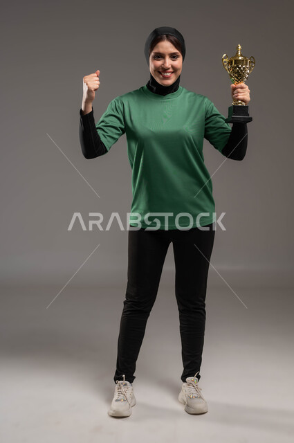 ممارسة رياضات وأنشطة بدنية ممتعة، المحافظة على نمط حياة صحي، بورتريه للاعبة عربية خليجية سعودية محجبة مبتسمة ترتدي الملابس الرياضية تحمل كأس البطولة وترفع يديها بإيماءات السعادة والفرح بالفور والانتصار، صورة لكامل الجسم خلفية رمادية.