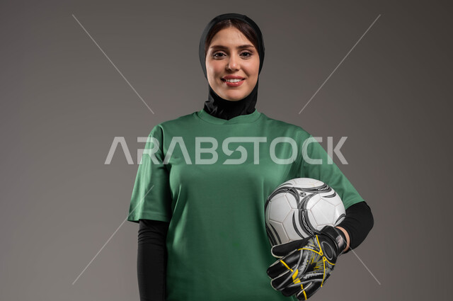 المراوغه وتسجيل الأهداف، نمط الحياة الصحية، أنشطة ترفيهية ورياضات نسائية، النظر إلى الكاميرا بإيماءات السرور، التدرب على اتقان لعب كرة القدم، بورتريه مقرب للاعبة عربية خليجية سعودية محجبة مبتسمة ترتدي ملابس رياضية تلعب دور حارس مرمى، خلفية رمادية