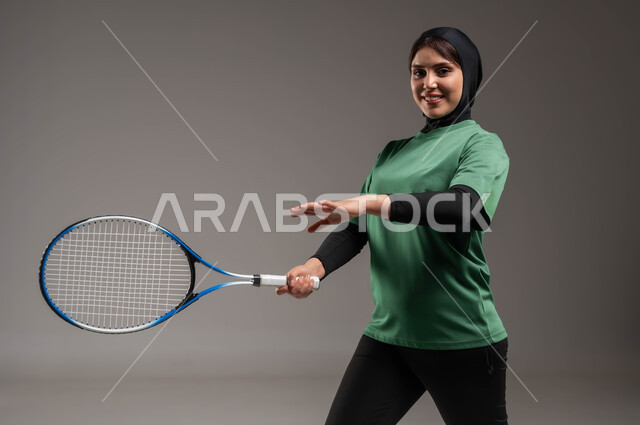 المحافظة على أسلوب الحياة الصحي، التدرب على إتقان لعبة البادل، أنشطة ترفيهية ورياضات نسائية، بورتريه للاعبة بادل عربية خليجية سعودية محجبة ترتدي الملابس الرياضية تمسك بيدها المضرب، النظر إلى الكاميرا إيماءات تدل على السعادة، خلفية رماديه