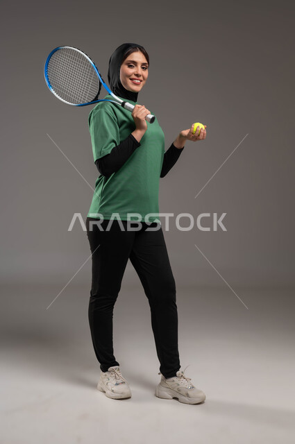 النظر إلى الكاميرا إيماءات تدل على السعادة، المحافظة على أسلوب الحياة الصحي، التدرب على إتقان لعبة البادل، أنشطة ترفيهية ورياضات نسائية، بورتريه للاعبة بادل عربية خليجية سعودية محجبة ترتدي الملابس الرياضية تمسك بيدها المضرب والكرة، صورة بكامل الجسم، خلفية رماديه