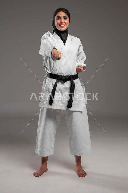 Enjoying the application of kata movements, women’s sports and exercises, performing combat movements specific to karate, a portrait of a veiled Saudi Gulf Arab young woman wearing a white sports uniform with a black belt, training in Japanese martial arts, a full-body portrait, gray background.