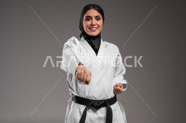 تطبيق حركات الكاتا, رياضات وتمارين نسائية، القيام بحركات قتالية خاصة برياضة الكاراتيه، بورتريه مقرب لشابة عربية خليجية سعودية محجبة ترتدي الزي الرياضي الأبيض مع الحزام الأسود تقوم بالتدرب على الفنون القتالية اليابانية بإيماءات المتعة والسعادة، خلفية رمادية