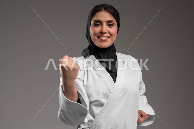 Women's sports and exercises, performing combat movements specific to karate, a close-up portrait of a veiled Saudi Gulf Arab young woman wearing a white sports uniform with a black belt, training in Japanese martial arts with gestures of pleasure and happiness, applying kata movements, gray background.