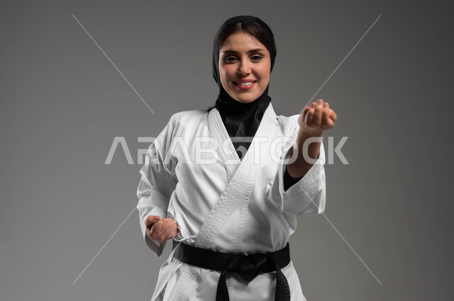 القيام بحركات قتالية خاصة برياضة الكاراتيه، بورتريه لشابة عربية خليجية سعودية محجبة ترتدي الزي الرياضي الأبيض مع الحزام الأسود تقوم بالتدرب على الفنون القتالية اليابانية، تطبيق حركات الكاتا, مفهوم اللياقة البدنية والتوازن، رياضات وتمارين نسائية، خلفية رمادية
