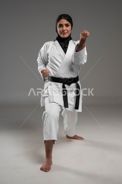 القيام بحركات قتالية خاصة برياضة الكاراتيه، التدرب على الفنون القتالية اليابانية, رياضات وتمارين نسائية، بورتريه لشابة عربية خليجية سعودية محجبة ترتدي الزي الرياضي الأبيض مع الحزام الأسود تقوم بتطبيق حركات الكاتا، صورة لكامل الجسم، خلفية رمادية