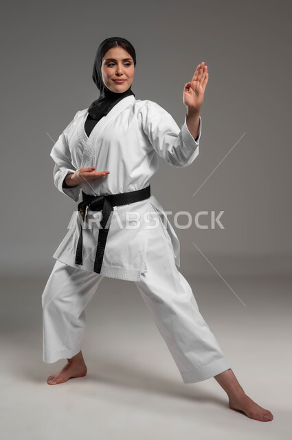 الاستعداد لممارسة رياضة الكاراتيه, التدرب على حركات قتالية خاصة, فنون الدفاع عن النفس, بورتريه لامرأة عربية سعودية خليجية ترتدي زي الكاراتيه والحجاب والحزام الاسود, صورة الجسم بالطول الكامل, خلفية رمادية