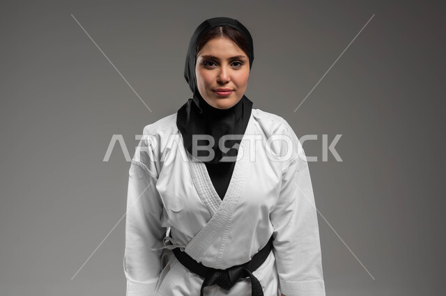 أداء تحية الكاراتيه, نادي رياضي نسائي في السعودية, بورتريه مقرب لامرأة عربية سعودية خليجية ترتدي زي الكاراتيه والحجاب والحزام الاسود تستعد للتدريب, أنشطة ترفيهية نسائية, خلفية رمادية