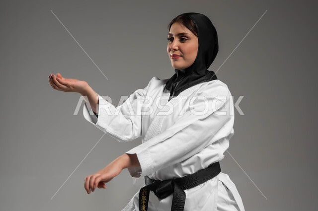 التمرن على حركات قتالية خاصة, مشاركة النساء السعوديات بالانشطة الرياضية ترفيهية, فنون الدفاع عن النفس, بورتريه مقرب لامرأة عربية سعودية خليجية ترتدي زي الكاراتيه  والحجاب والحزام الاسود ترفع يدها للاستعداد للتدريب, خلفية رمادية