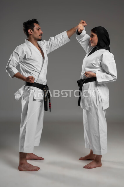 التمرن على الحركات القتالية, بطولات رياضة الكاراتيه, بورتريه لبنت وشاب عربيان سعوديان خليجيان يرتديان زي الكاراتيه الابيض والحزام الاسود وترتدي الشابة الحجاب الاسود يقومان بالتدريب على البطولة, صورة الجسم بالطول الكامل, خلفية رمادية