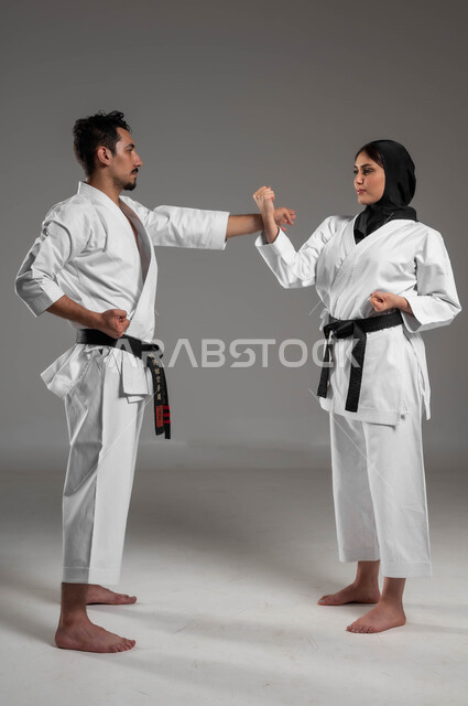 التعليم وتطوير المهارات الحركية, ايماءات التركيز والانتباه, القيام بحركات قتالية خاصة والدفاع عن النفس, بورتريه من الجانب لبنت وشاب عربيان سعوديان خليجيان يرتديان زي الكارتيه الابيض والحزام الاسود , الاستعداد لصد الضربات, صورة الجسم بالطول الكامل, خلفية رمادية