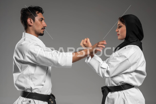 القيام بحركات الدفاع عن النفس, التعليم وتطوير المهارات الحركية, بورتريه مقرب من الجانب لبنت وشاب عربيان سعوديان خليجيان يرتديان زي الكارتيه الابيض والحزام الاسود يقومان بالتدريب على البطولة, مشاركة النساء السعوديات بالبطولات والأنشطة الرياضية, خلفية رمادية