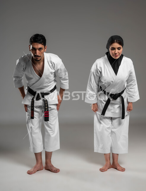 مشاركة النساء السعوديات في الرياضات القتالية, النظر إلى الكاميرا والاستعداد للمواجهة, ايماءات التحدي والحزم, بورتريه لبنت وشاب عربيان سعوديان خليجيان يلبسان لباس الكارتبه الابيض والحزام الاسود يقومان بأداء تحية الكاراتيه, صورة الجسم بالطول الكامل, خلفية رمادية