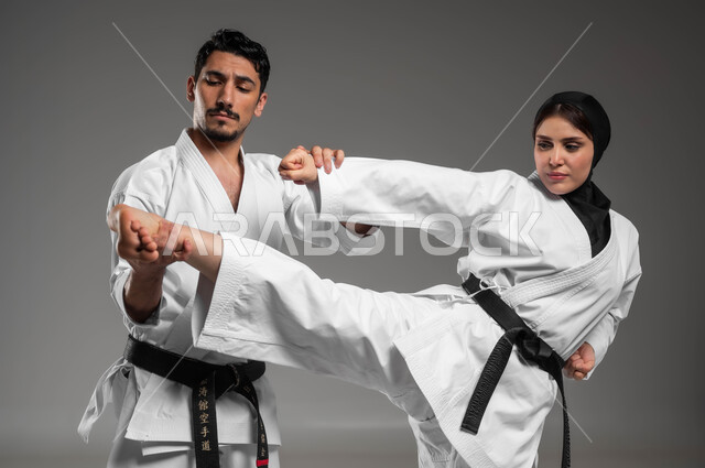 التمرن على الحركات القتالية, التعليم وتطوير المهارات الحركية, بطولات رياضية الكارتيه, بورتريه لبنت وشاب عربيان سعوديان خليجيان يرتديان زي الكارتيه الابيض والحزام الاسود يقومان بالتدريب على البطولة, رياضة الألعاب الأولمبية, خلفية رمادية