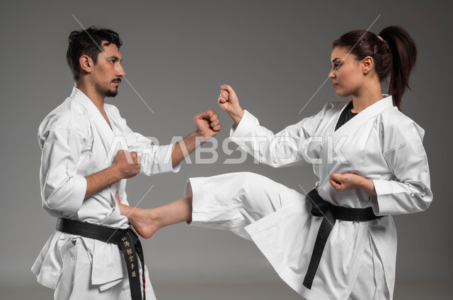 التعليم وتطوير المهارات الحركية, بطولات وأنشطة رياضة الكاراتيه, التمرن على الحركات القتالية, بورتريه لبنت وشاب عربيان سعوديان خليجيان يرتديان زي الكاراتيه الابيض والحزام الاسود يقومان بالتدريب على البطولة, الألعاب الأولمبية السعودية, خلفية رمادية