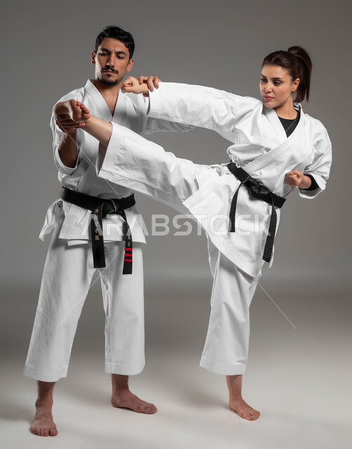 ايماءات التركيز والتحدي, بطولات رياضية الكارتيه, التعليم وتطوير المهارات, بورتريه لبنت وشاب عربيان سعوديان خليجيان يرتديان زي الكارتيه الابيض والحزام الاسود, القيام بحركات قتالية والدفاع عن النفس, صورة الجسم بالطول الكامل, خلفية رمادية