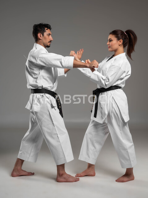 ايماءات التركيز والانتباه, القيام بحركات قتالية خاصة والدفاع عن النفس, التعليم وتطوير المهارات الحركية, بورتريه من الجانب لبنت وشاب عربيان سعوديان خليجيان يرتديان زي الكارتيه الابيض والحزام الاسود, الحفاظ على اللياقة البدنية, صورة الجسم بالكامل, خلفية رمادية