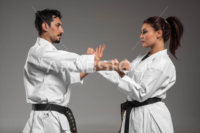 القيام بحركات قتالية خاصة والدفاع عن النفس, التعليم وتطوير المهارات الحركية, بورتريه مقرب من الجانب لبنت وشاب عربيان سعوديان خليجيان يرتديان زي الكارتيه الابيض والحزام الاسود, الحفاظ على اللياقة البدنية, خلفية رمادية