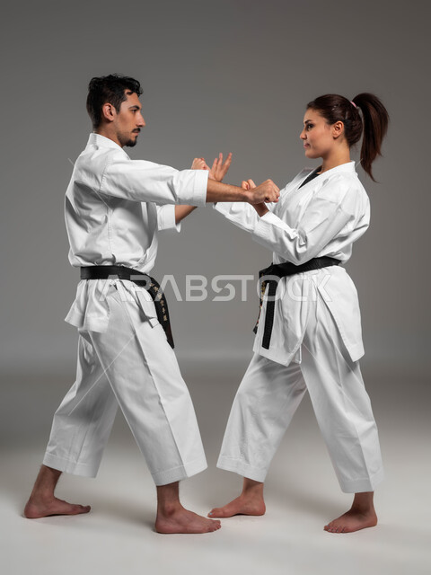 التعليم وتطوير المهارات الحركية, القيام بحركات قتالية خاصة والدفاع عن النفس, بورتريه من الجانب لبنت وشاب عربيان سعوديان خليجيان يرتديان زي الكارتيه الابيض والحزام الاسود, الحفاظ على اللياقة البدنية, صورة الجسم بالطول الكامل, خلفية رمادية