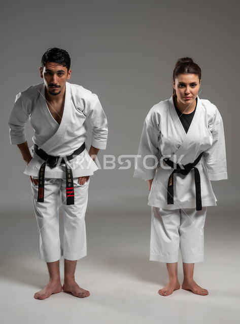 النظر إلى الكاميرا والاستعداد الى المواجهة, ايماءات التحدي والحزم, بورتريه لبنت وشاب عربيان سعوديان خليجيان يرتديان لباس الكاراتيه الابيض والحزام الاسود, أداء تحية الكاراتييه, صورة الجسم بالطول الكامل, خلفية رمادية