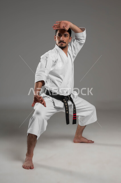 تطبيق خطوات الكاتا، أسلوب حياة صحي، ممارسة فنون قتال يابانية، مفهوم اللياقة البدنية، التدرب على حركات قتال خاصة وفن الدفاع عن النفس, بورتريه لرجل عربي خليجي سعودي يرتدي الزي الرياضي الأبيض مع حزام أسود يمارس الكاراتيه، صورة لكامل الجسم، خلفية رمادية
