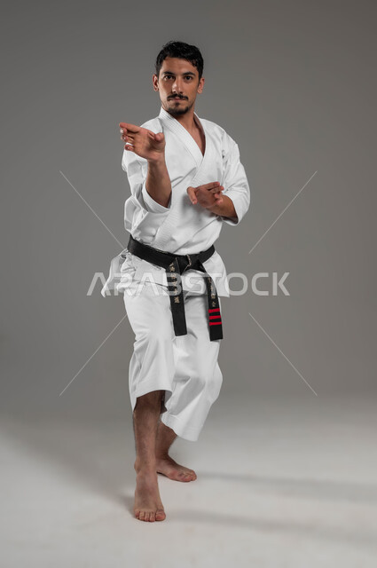 النظر إلى الكاميرا بثقة، فنون قتال يابانية، تطبيق خطوات الكاتا، التدرب على حركات قتالية خاصة وفن الدفاع عن النفس, بورتريه  لرجل عربي خليجي سعودي يرتدي الزي الرياضي الأبيض مع حزام أسود يمارس الكاراتيه بإيماءات التركيز والانتباه، صورة لكامل الجسم، خلفية رمادية