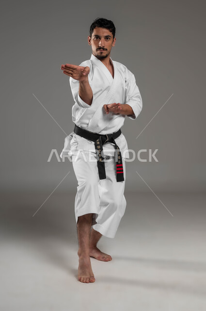الاستعداد لبدء القتال، ممارسة فنون قتال يابانية، مفهوم اللياقة البدنية، تطبيق خطوات الكاتا، التدرب على حركات قتال خاصة وفن الدفاع عن النفس, بورتريه لرجل عربي خليجي سعودي يرتدي الزي الرياضي الأبيض مع حزام أسود يمارس الكاراتيه، صورة لكامل الجسم، خلفية رمادية