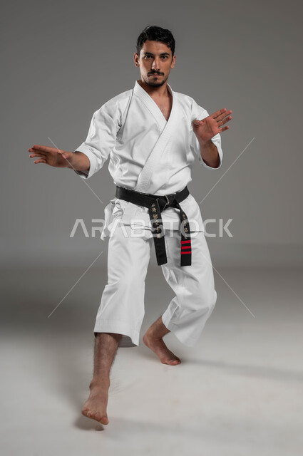 أسلوب حياة صحي، ممارسة فنون قتال يابانية، مفهوم اللياقة البدنية، تطبيق خطوات الكاتا، التدرب على حركات قتال خاصة وفن الدفاع عن النفس, بورتريه لرجل عربي خليجي سعودي يرتدي الزي الرياضي الأبيض مع حزام أسود يمارس الكاراتيه، صورة لكامل الجسم، خلفية رمادية
