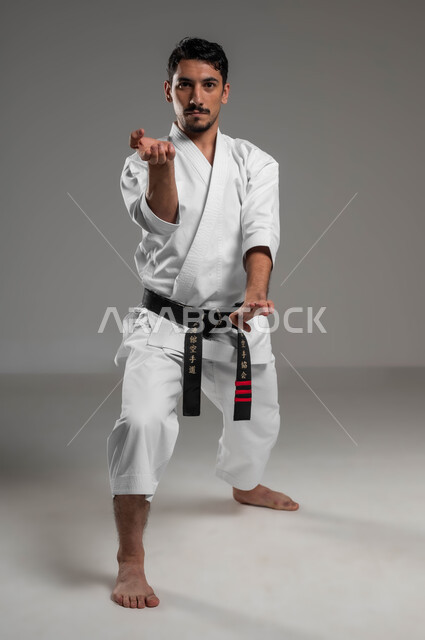 التركيز و الانتباه أثناء ممارسة فنون قتال يابانية، مفهوم اللياقة البدنية، تطبيق خطوات الكاتا، التدرب على حركات قتال خاصة وفن الدفاع عن النفس, بورتريه لرجل عربي خليجي سعودي يرتدي الزي الرياضي الأبيض مع حزام أسود يمارس الكاراتيه، صورة لكامل الجسم، خلفية رمادية
