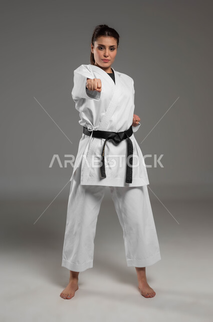 النظر إلى الكاميرا بثقة، فنون قتال يابانية، تطبيق خطوات الكاتا، التدرب على حركات قتالية خاصة وفن الدفاع عن النفس, بورتريه  لفتاة عربية خليجية سعودية ترتدي الزي الرياضي الأبيض مع حزام أسود تمارس الكاراتيه بإيماءات التركيز والانتباه، صورة لكامل الجسم، خلفية رمادية
