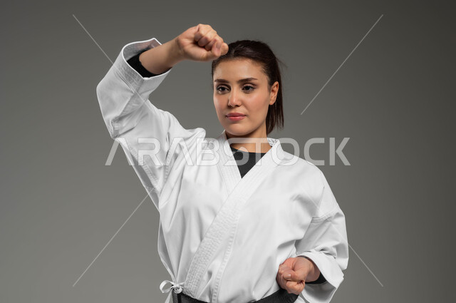 ممارسة فن الدفاع عن النفس وتعلم فنون قتالية يابانية، أنشطة ورياضات تنافسية، تطبيق خطوات الكاتا، تنفيذ تمارين رياضية للمحافظة على اللياقة، بورتريه مقرب لفتاة عربية خليجية سعودية ترتدي الزي الرياضي الأبيض مع حزام أسود تمارس الكاراتيه بتعابير التركيز، خلفية رمادية