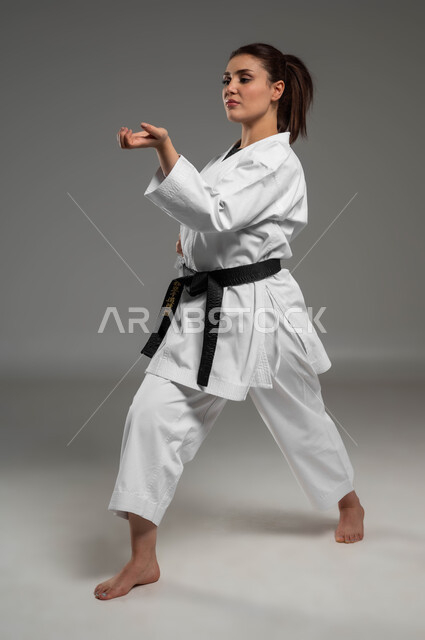 تعلم فنون قتال يابانية وتطبيق خطوات الكاتا، التدرب على حركات قتالية خاصة وفن الدفاع عن النفس, بورتريه  لفتاة عربية خليجية سعودية ترتدي الزي الرياضي الأبيض مع حزام أسود تمارس الكاراتيه بإيماءات التركيز والانتباه، صورة لكامل الجسم، خلفية رمادية