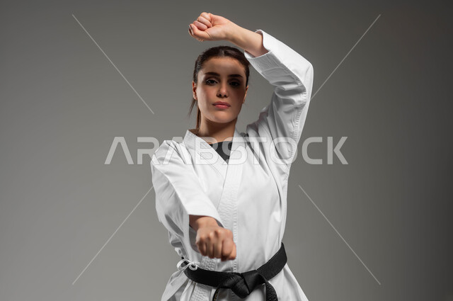 جودة الحياة الصحية، ممارسة فن الدفاع عن النفس وتعلم فنون قتالية يابانية، تطبيق خطوات الكاتا، تنفيذ تمارين رياضية للمحافظة على اللياقة، بورتريه مقرب لفتاة عربية خليجية سعودية ترتدي الزي الرياضي الأبيض مع حزام أسود تمارس الكاراتيه بتعابير التركيز، خلفية رمادية