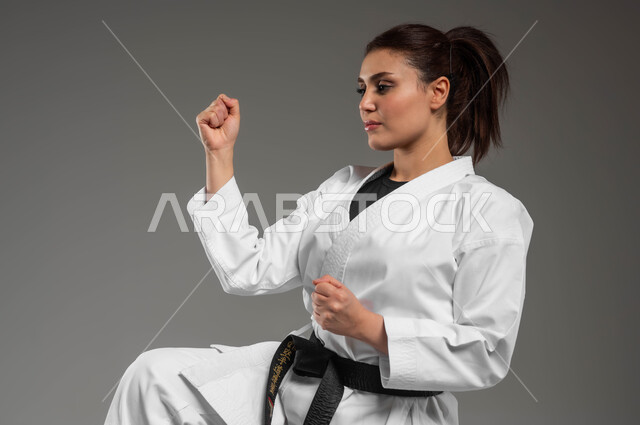 الاستعداد لتوجيه ضربة، تطبيق خطوات الكاتا، تنفيذ تمارين رياضية للمحافظة على اللياقة، فن الدفاع عن النفس, تعلم فنون قتال يابانية، بورتريه مقرب لفتاة عربية خليجية سعودية ترتدي الزي الرياضي الأبيض مع حزام أسود تمارس الكاراتيه بتعابير التركيز، خلفية رمادية